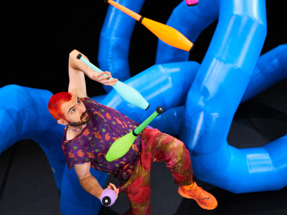 Ein Mann mit roten Haaren jongliert auf der Bühne mit bunten Keulen vor großen, sich windenden blauen Ballonfiguren.