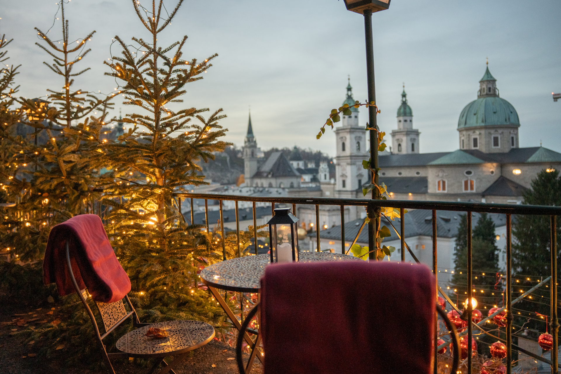 Gemütlicher Balkon mit festlicher Beleuchtung, kleinem Tisch und Blick auf die Stadt mit Kirchenkuppeln in der Abenddämmerung.