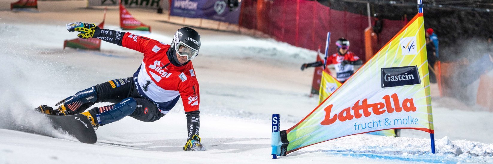 Ein Snowboarder in einem roten Trikot dreht sich während eines Snowboardrennens auf einer verschneiten Piste scharf um eine Fahne.