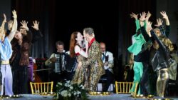 Jedermann und die Buhlschaft umarmen sich mitten auf der Bühne, während Musiker spielen und Tänzer mit erhobenen Armen sie an einem festlich geschmückten Tisch umringen.