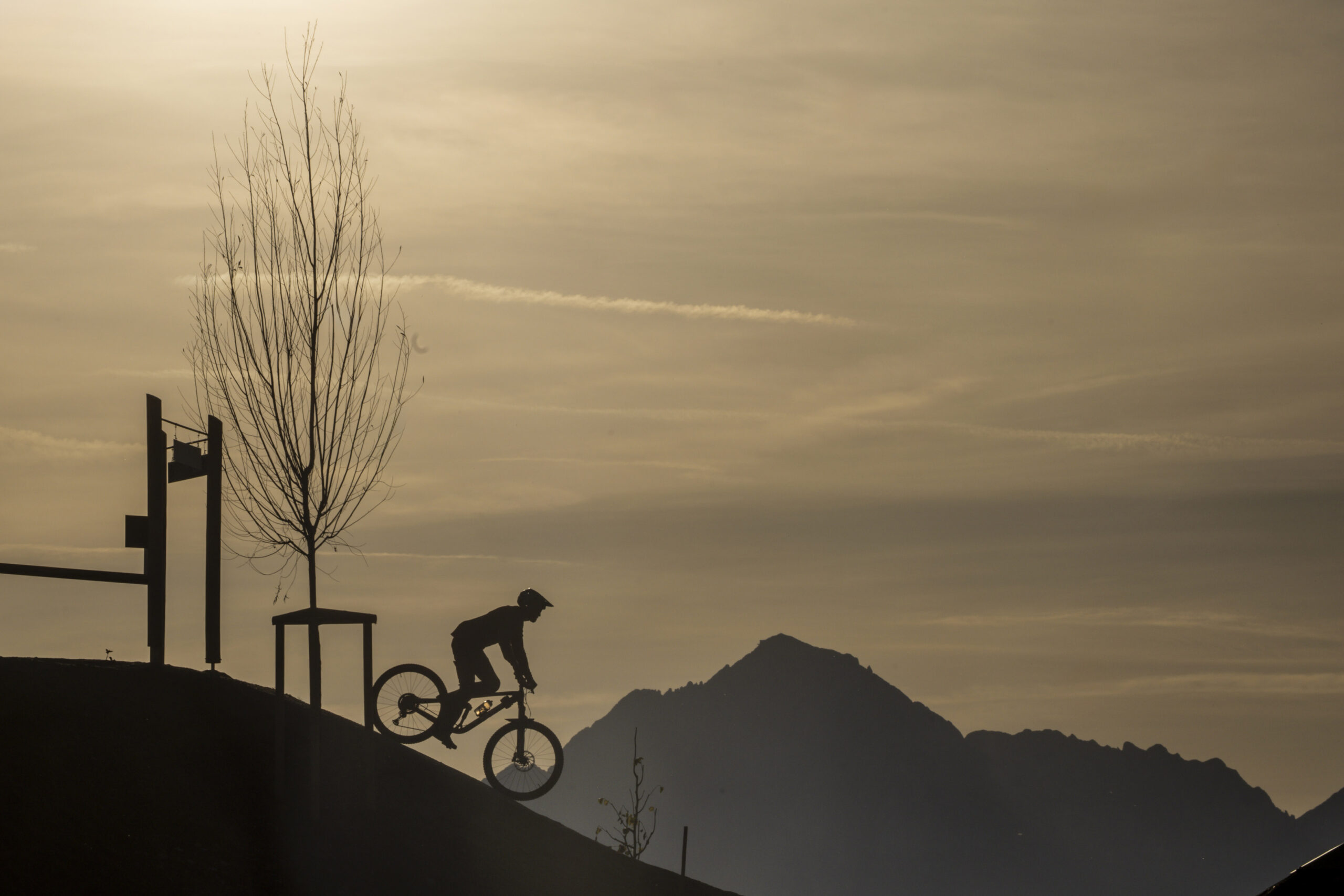 Silhouette einer Person, die mit dem Fahrrad einen Hügel hinauffährt, mit Bergen und einem Baum im Hintergrund bei Sonnenuntergang.