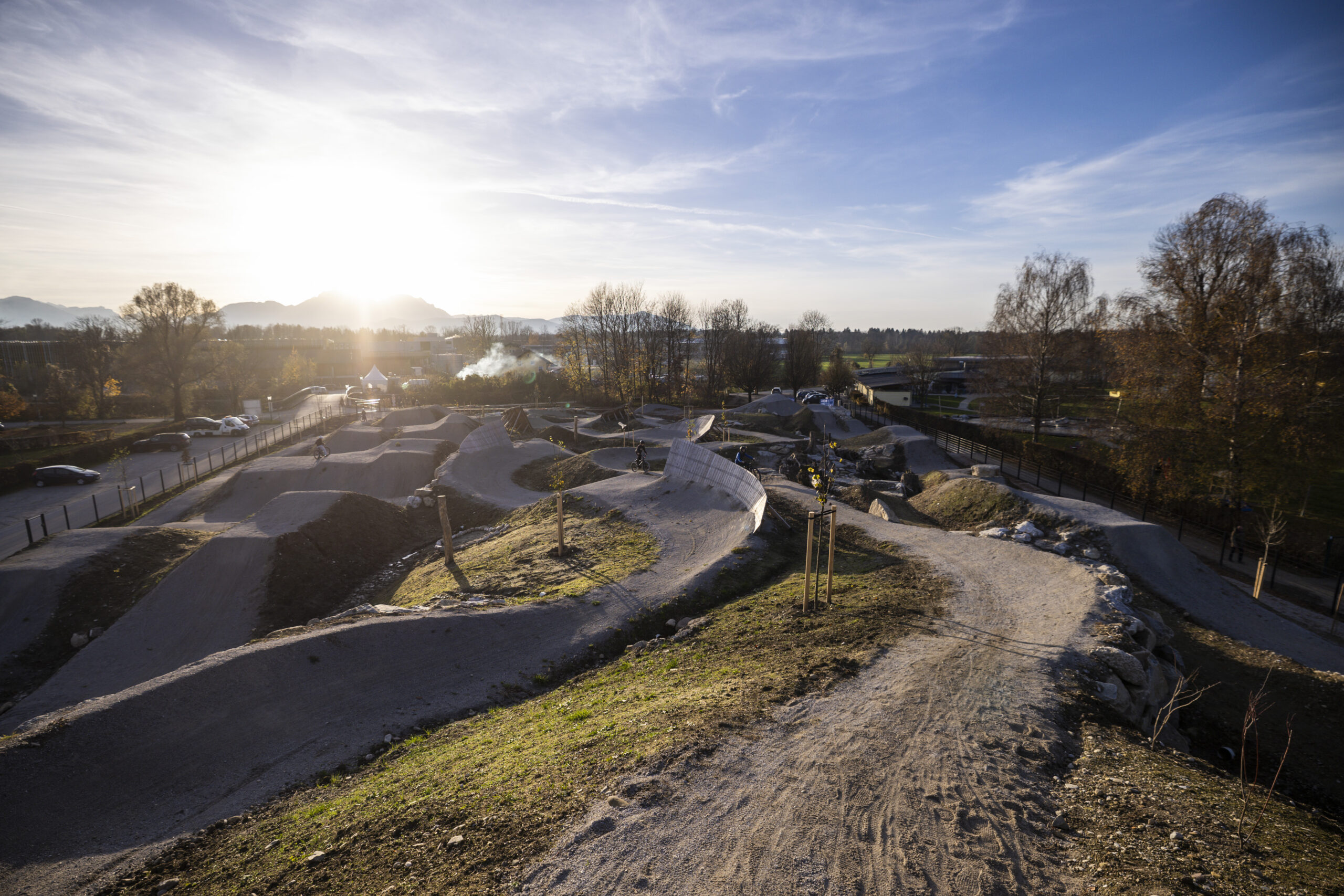 Ein gewundener unbefestigter Fahrradweg in einem Park bei Sonnenuntergang, mit Bäumen und Autos im Hintergrund.