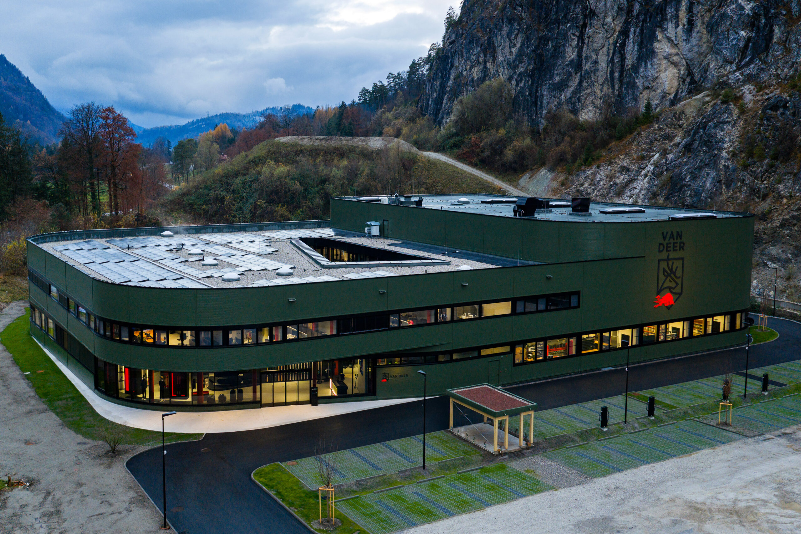 Modernes grünes Gebäude mit großen Fenstern vor einem felsigen Hügel und bewölktem Himmel.