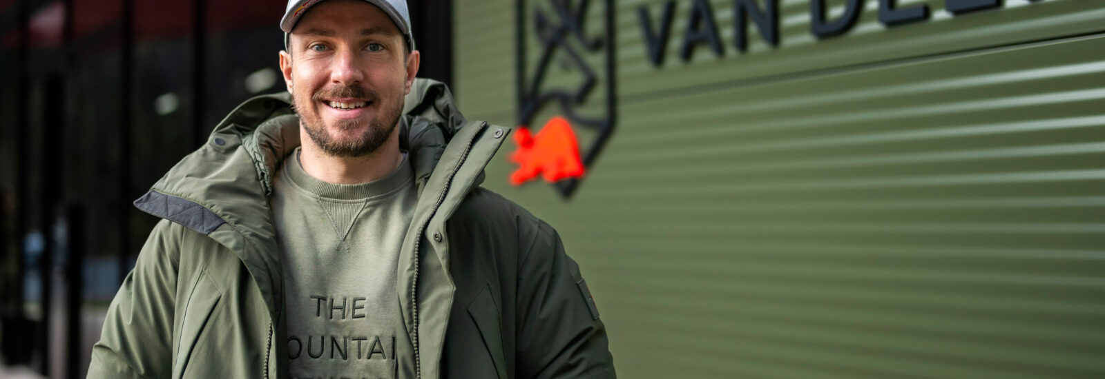 Ein lächelnder Mann in grüner Jacke und Mütze steht vor einem Gebäude mit dem VAN DEER-Logo an einer grünen Wand.