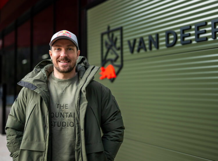 Ein lächelnder Mann in grüner Jacke und Mütze steht vor einem Gebäude mit dem VAN DEER-Logo an einer grünen Wand.