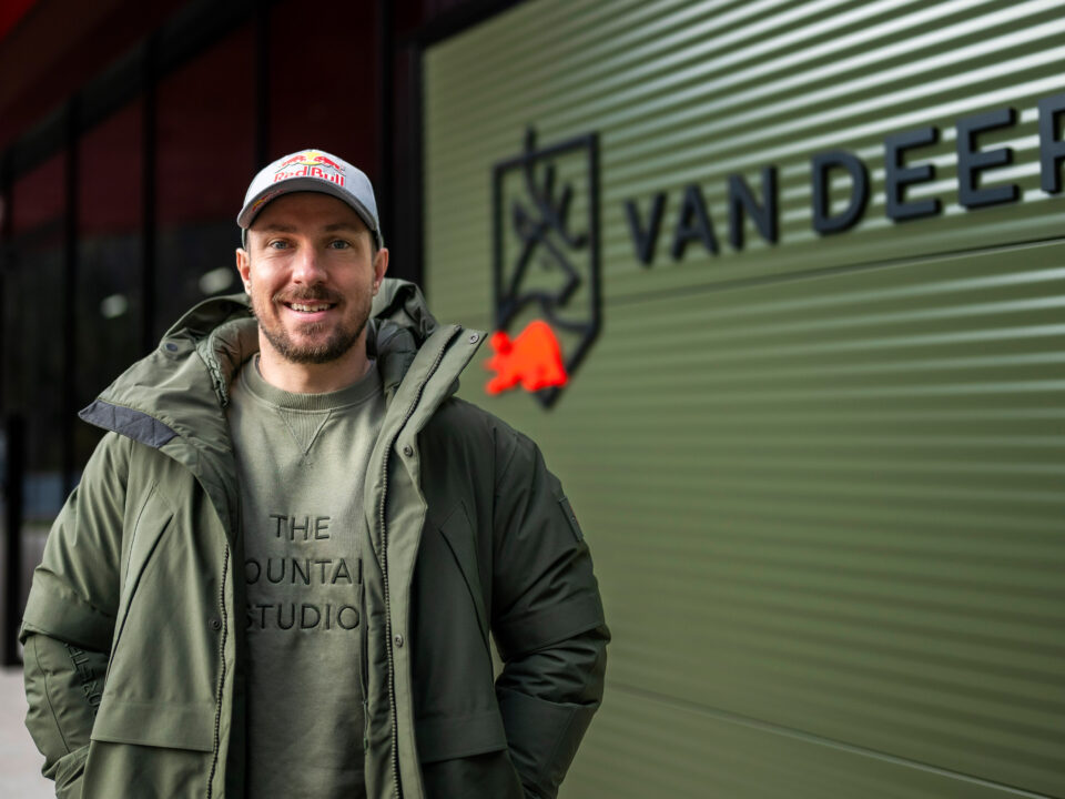 Ein lächelnder Mann in grüner Jacke und Mütze steht vor einem Gebäude mit dem VAN DEER-Logo an einer grünen Wand.