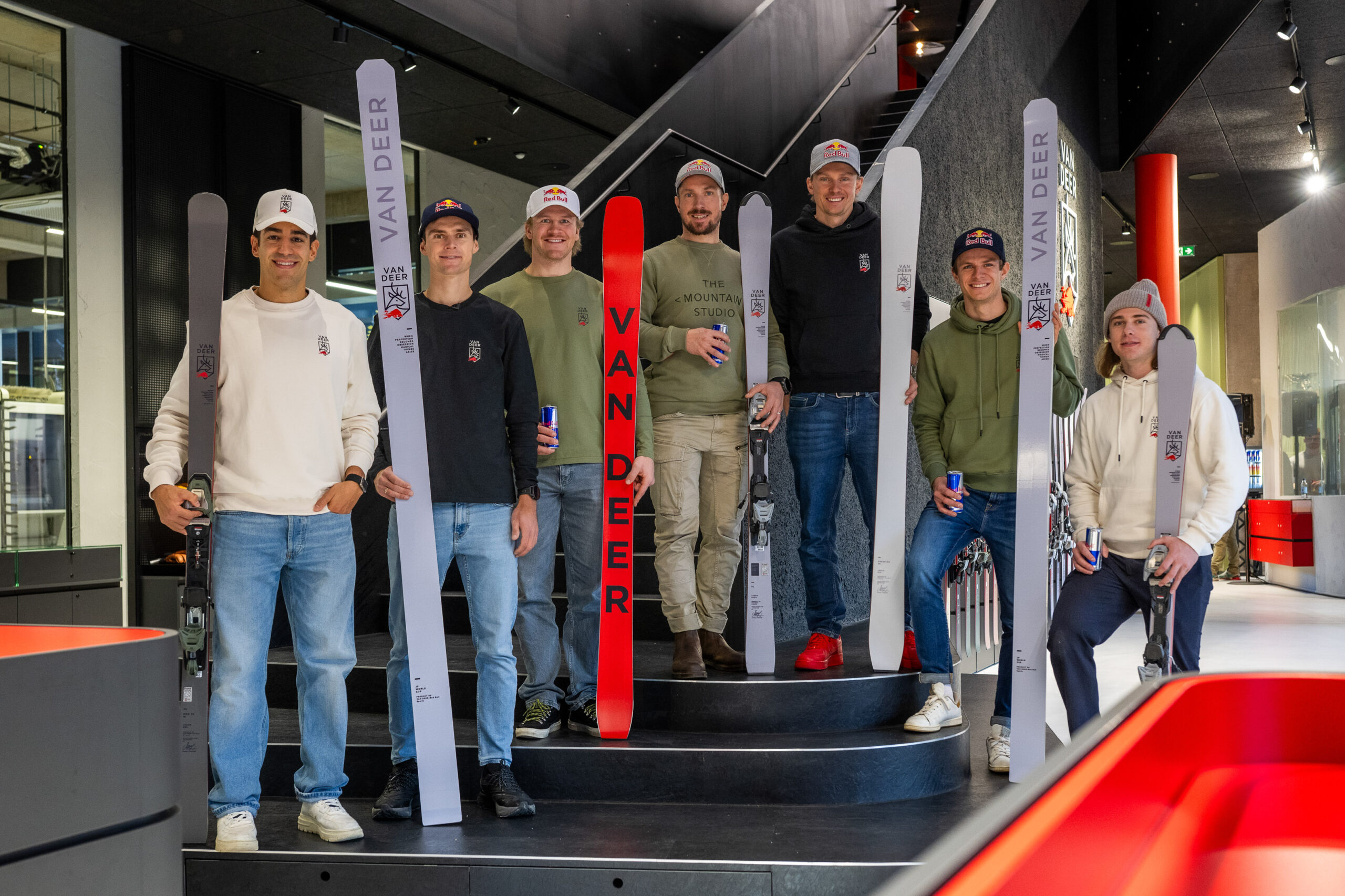 Sieben Personen stehen auf einer Treppe im Haus, jede hält Skier mit der Aufschrift Van Deer und lächelt in die Kamera.
