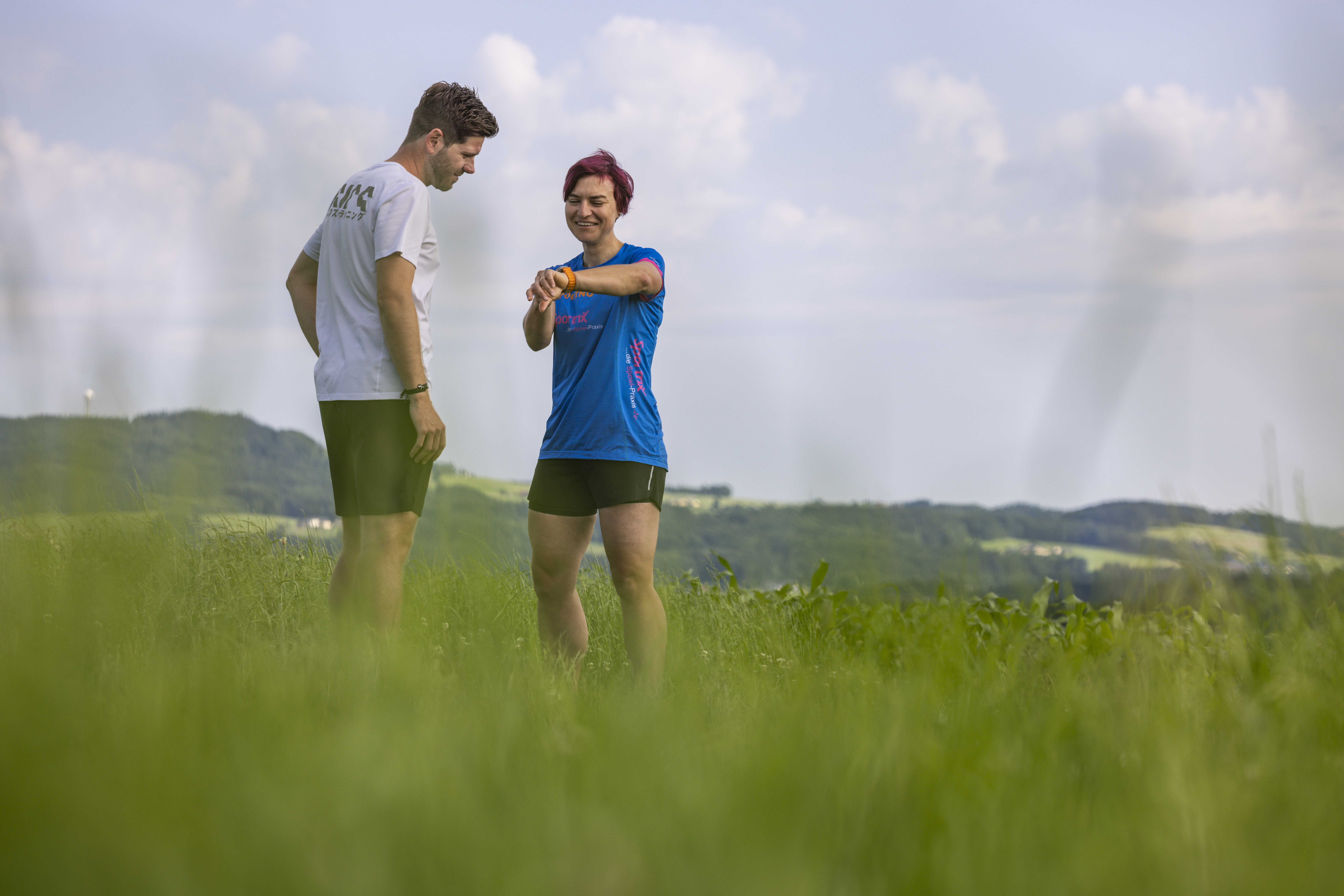 Zwei Personen in Sportkleidung geben Tipps zum Thema Herzgesundheit und Training, während sie auf einer Wiese eine Smartwatch überprüfen.