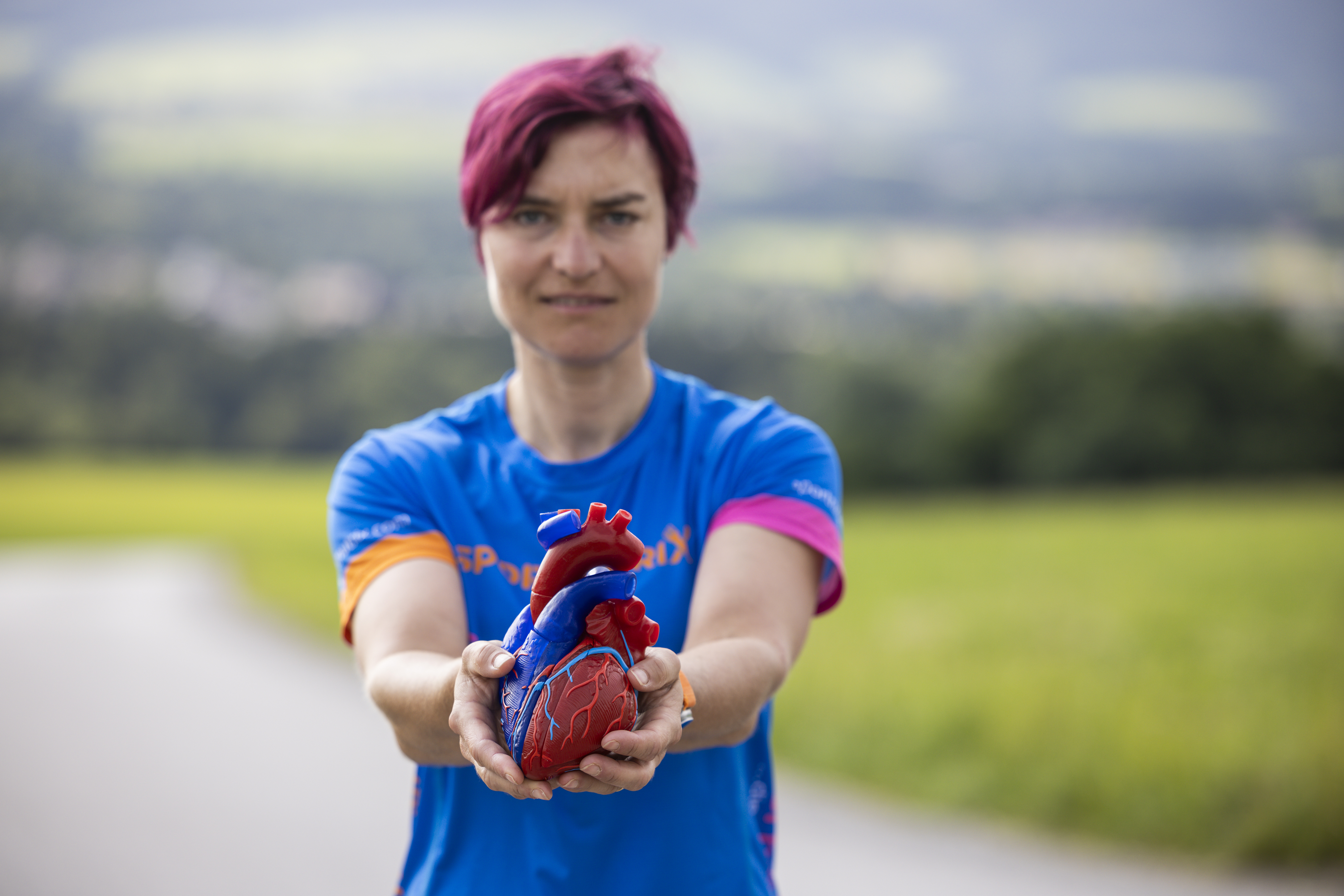 Person mit kurzen rosa Haaren hält ein Plastikherz und gibt Tipps zum Thema Herzgesundheit und Training im Freien.