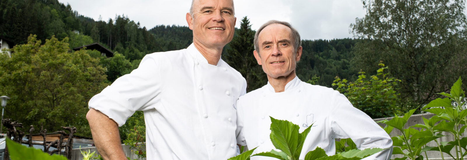 Zwei Köche in weißen Uniformen stehen zusammen im Freien zwischen grünen Pflanzen und Bäumen und lächeln in die Kamera.