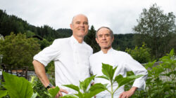 Zwei Köche in weißen Uniformen stehen zusammen im Freien zwischen grünen Pflanzen und Bäumen und lächeln in die Kamera.