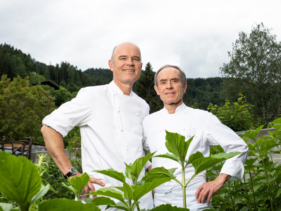Zwei Köche in weißen Uniformen stehen zusammen im Freien zwischen grünen Pflanzen und Bäumen und lächeln in die Kamera.