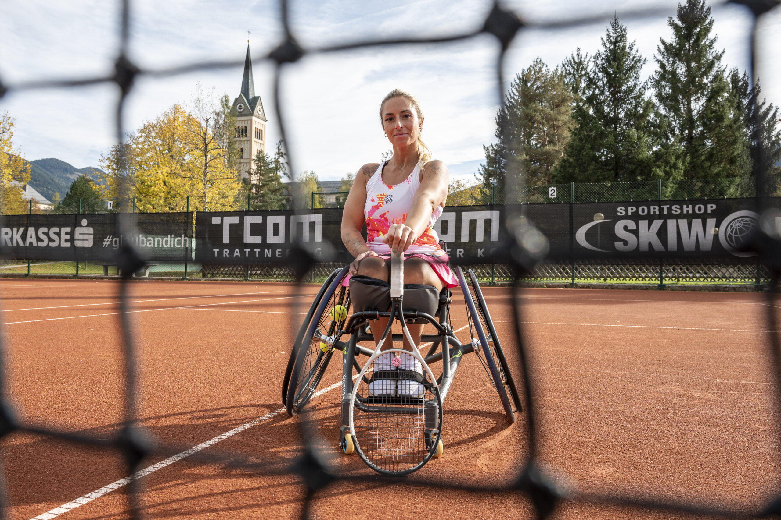 Eine Frau im Rollstuhl hält einen Tennisschläger auf einem Sandplatz, durch das Netz gesehen.