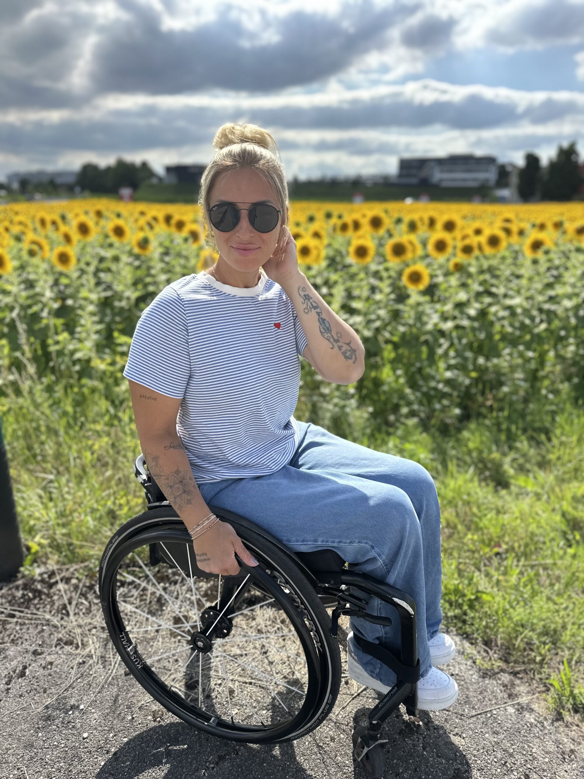 Eine Frau im Rollstuhl posiert an einem sonnigen Tag vor einem Sonnenblumenfeld, trägt eine Sonnenbrille und legere Kleidung.