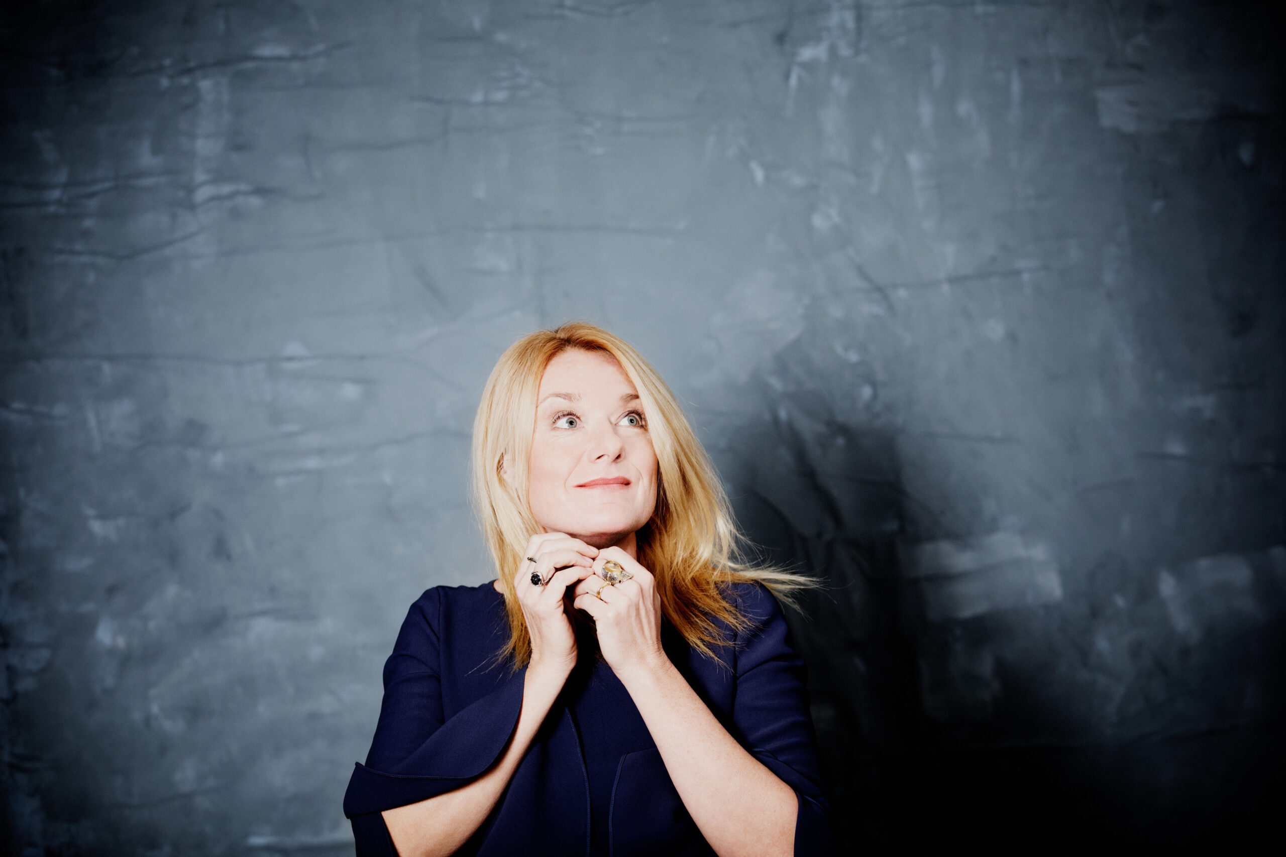 Eine blonde Frau in einem marineblauen Oberteil blickt nachdenklich vor einem strukturierten grauen Hintergrund auf.