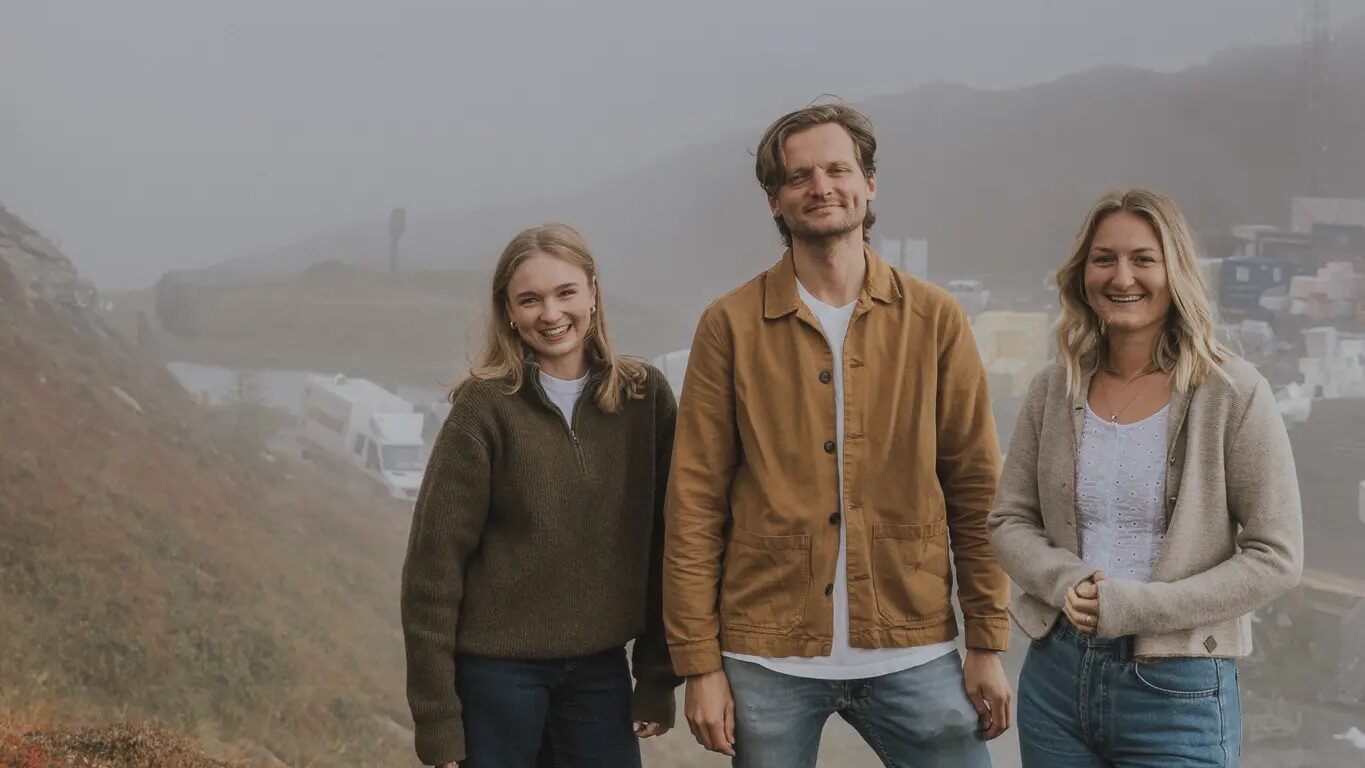 Drei Personen stehen lächelnd auf einem grasbewachsenen Hügel mit nebligem Wetter und geparkten Autos im Hintergrund.
