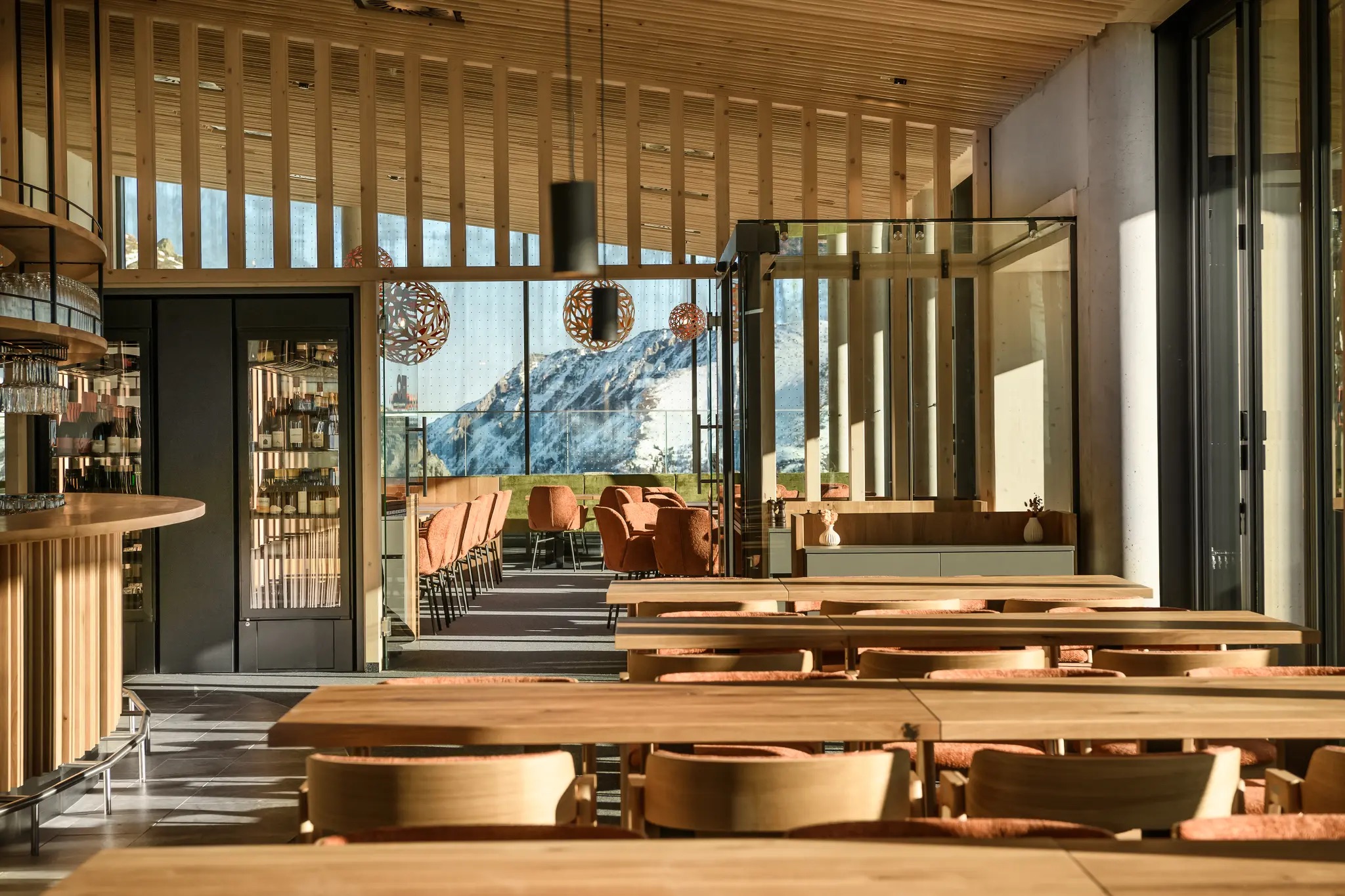 Modernes Restaurantinterieur mit Holztischen und -stühlen, großen Fenstern und Bergblick im Hintergrund.
