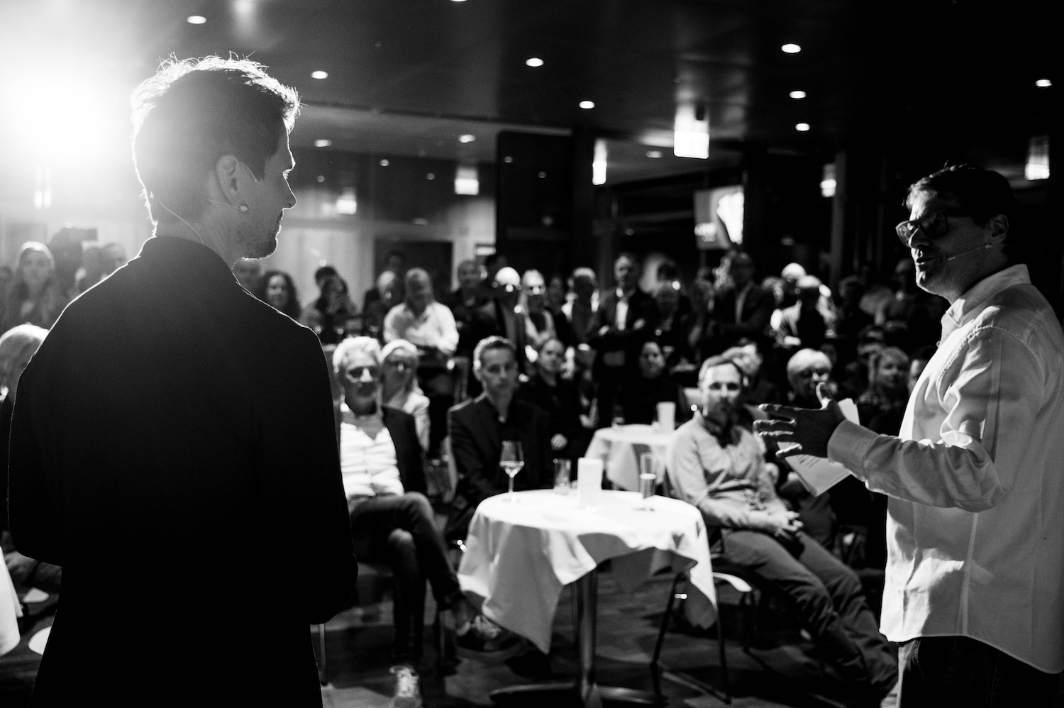 Zwei Personen sprechen zu einem aufmerksamen Publikum in einem dunklen, überfüllten Raum mit runden Tischen, in schwarz-weiß.