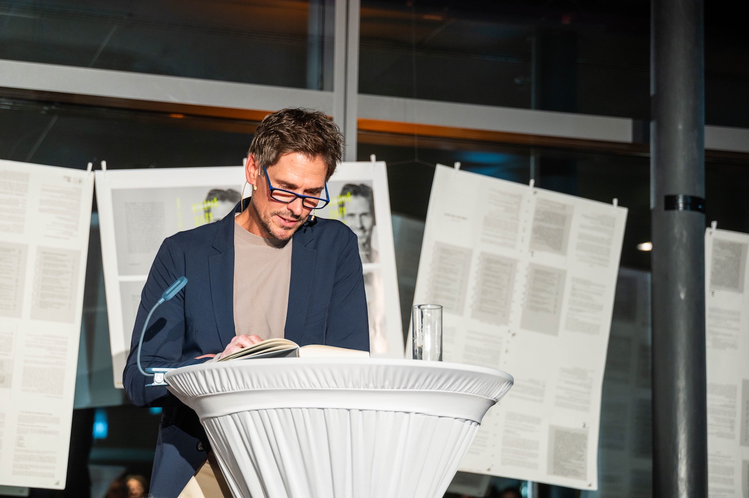 Ein Mann liest auf einem weißen Podium aus einem Buch, während im Hintergrund Dokumente und Fotos zu sehen sind.