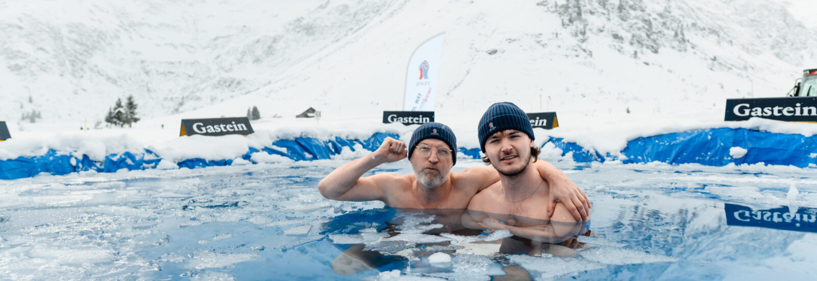 Zwei Männer mit Strickmützen posieren zusammen in einem eisigen Freibad, umgeben von schneebedeckten Bergen.