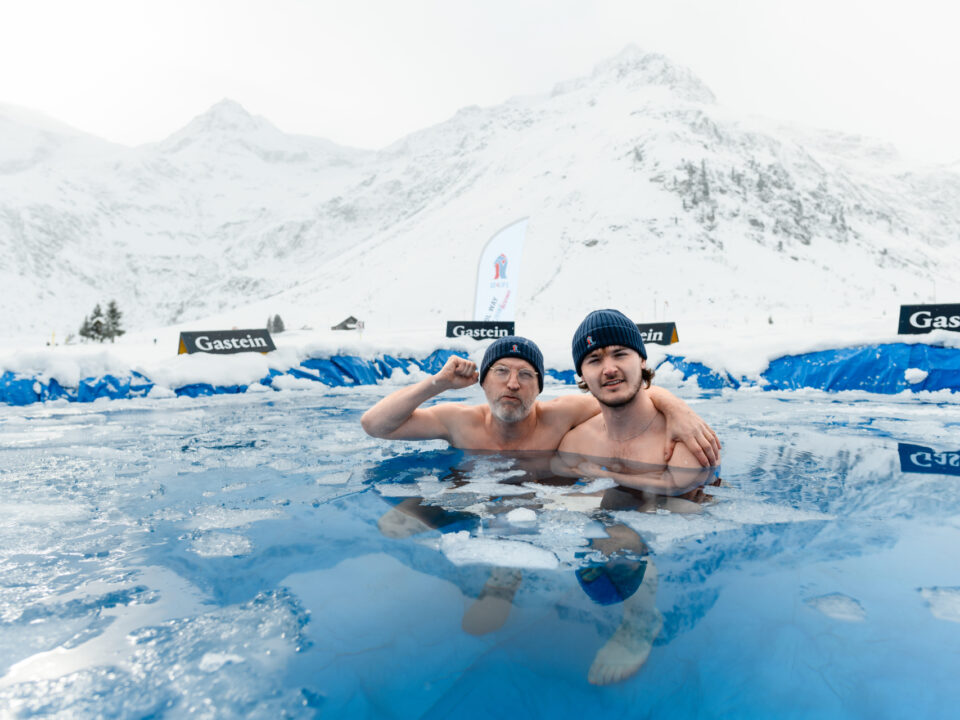 Zwei Männer mit Strickmützen posieren zusammen in einem eisigen Freibad, umgeben von schneebedeckten Bergen.