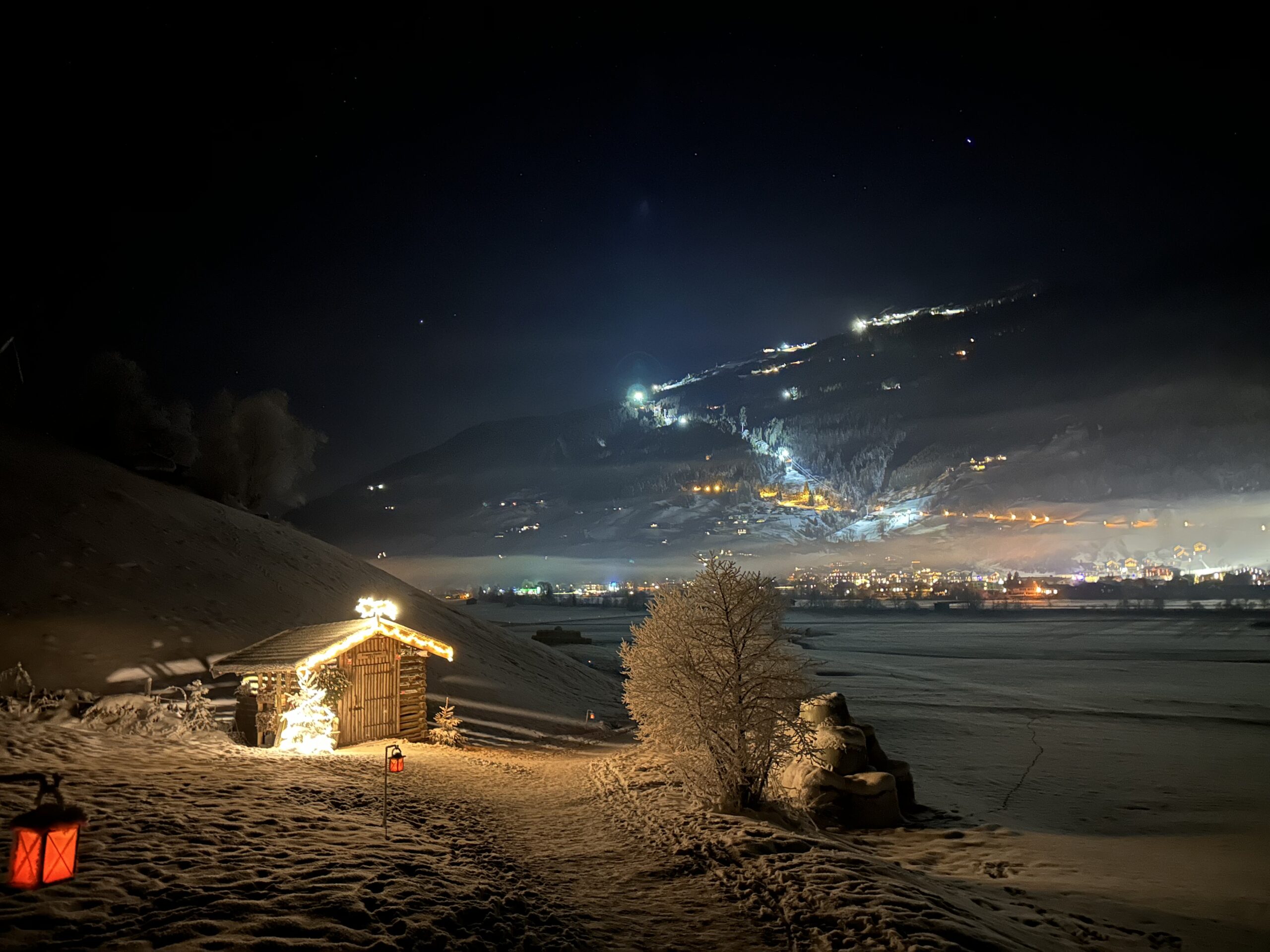Verschneite Hütte mit Weihnachtsbeleuchtung bei Nacht am Laternenweg Bramberg, mit Blick auf eine leuchtende Stadt und Berge.