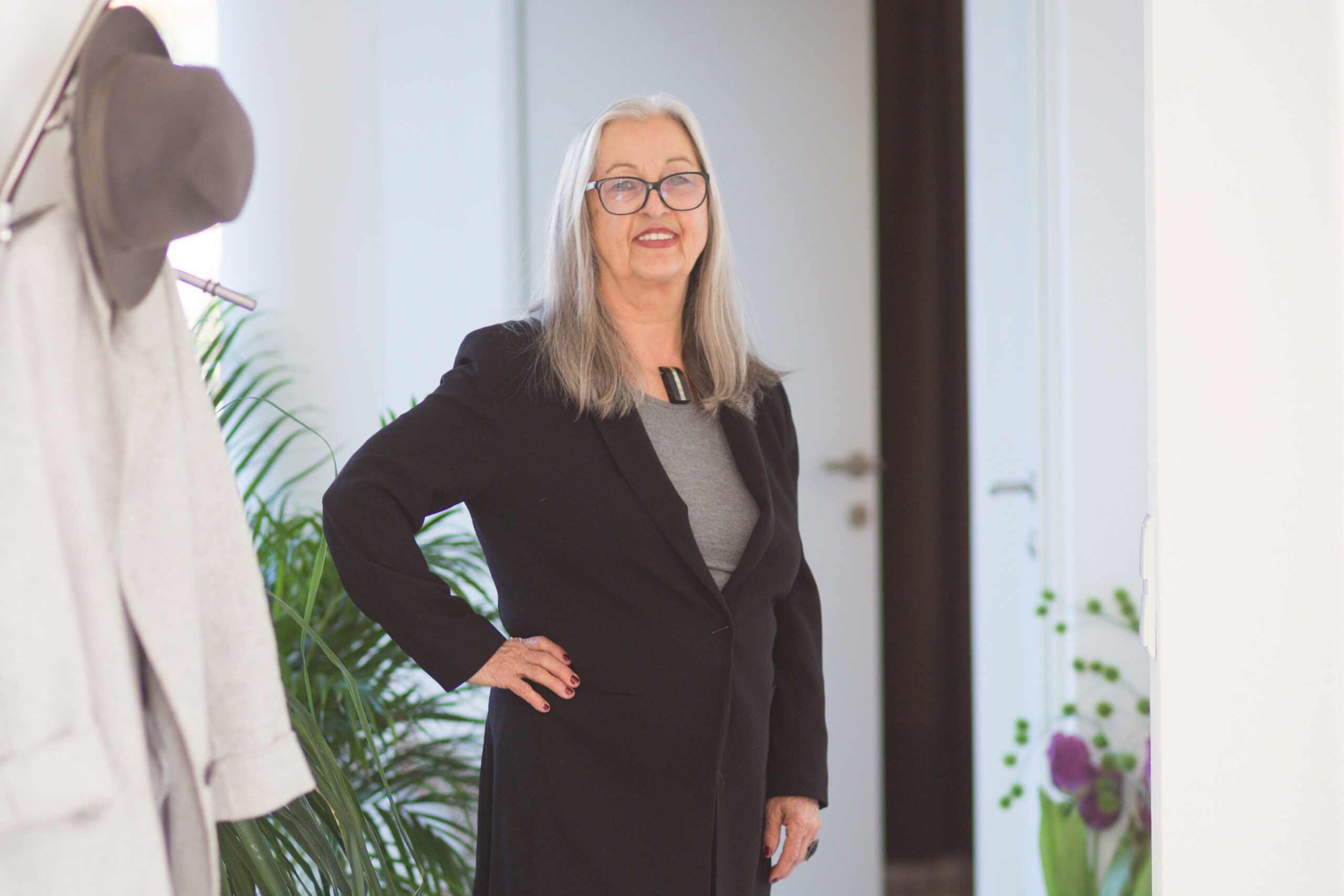 Ältere Frau mit grauen Haaren in einem schwarzen Blazer, lächelnd in einem Innenraum in der Nähe von Pflanzen bei Marias Biokosmetik Salzburg.