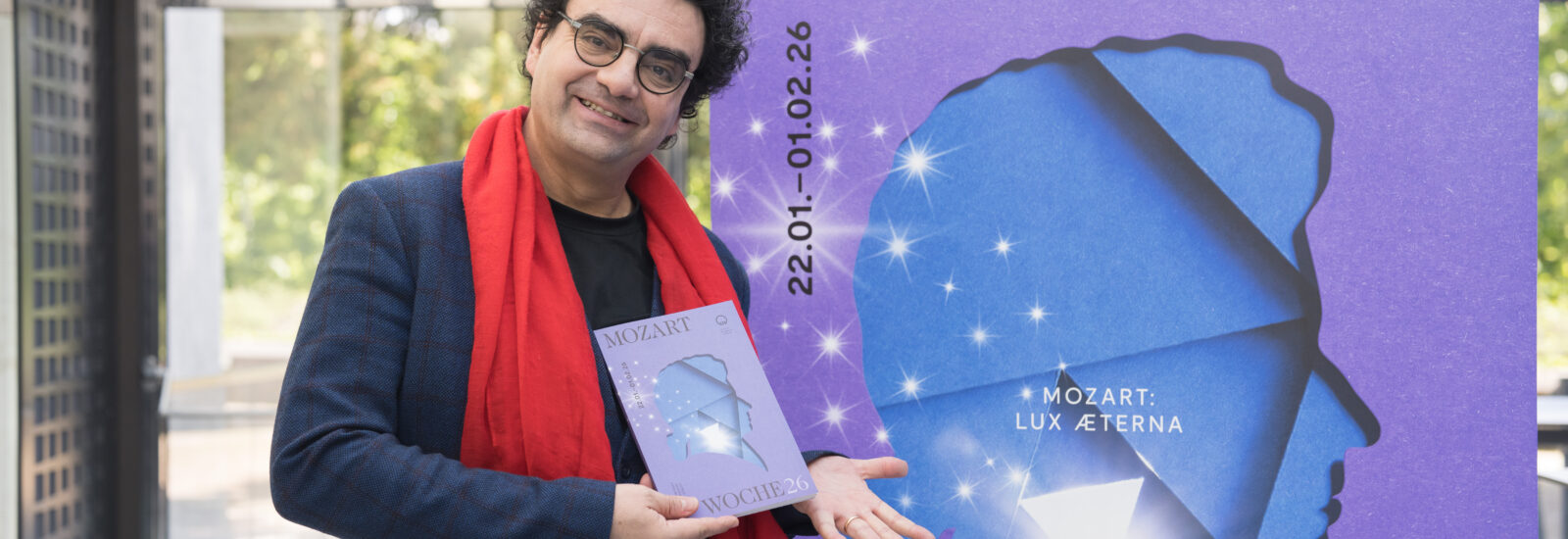 Ein lächelnder Mann mit rotem Schal hält ein Büchlein neben einem großen lila Mozart: Lux Aeterna-Plakat.