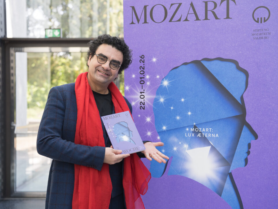 Ein lächelnder Mann mit rotem Schal hält ein Büchlein neben einem großen lila Mozart: Lux Aeterna-Plakat.