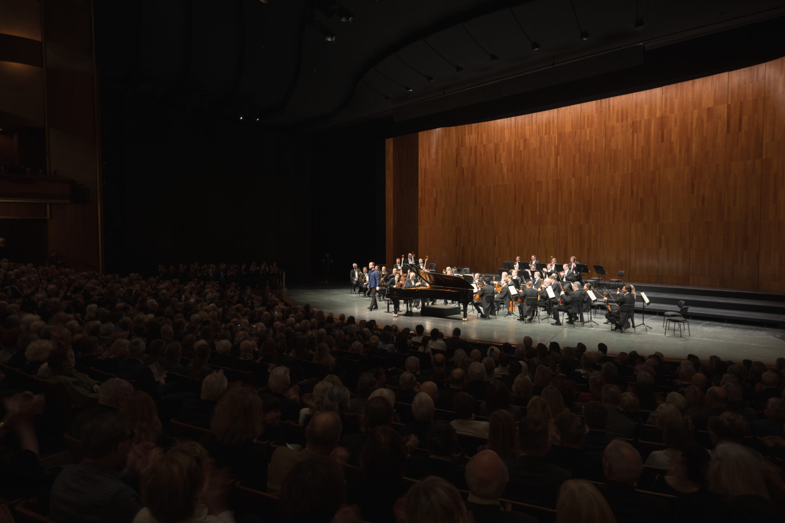 Ein Sinfonieorchester spielt auf der Bühne mit einem Flügel vor einem großen, sitzenden Publikum in einem Konzertsaal.