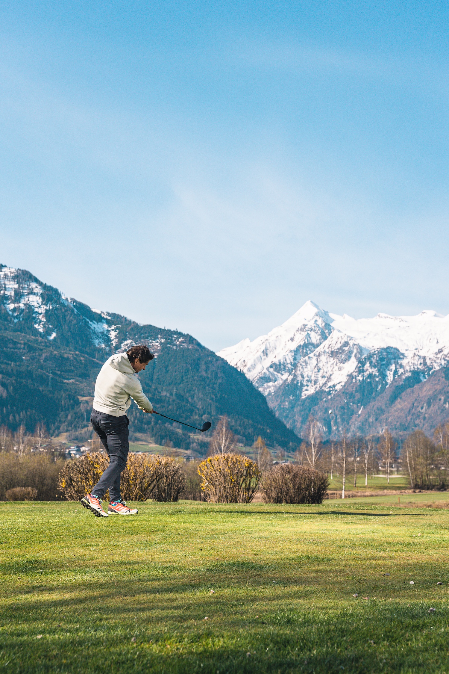 Eine Person schwingt einen Golfschl&auml;ger auf einer gr&uuml;nen Wiese mit verschneiten Bergen im Hintergrund.