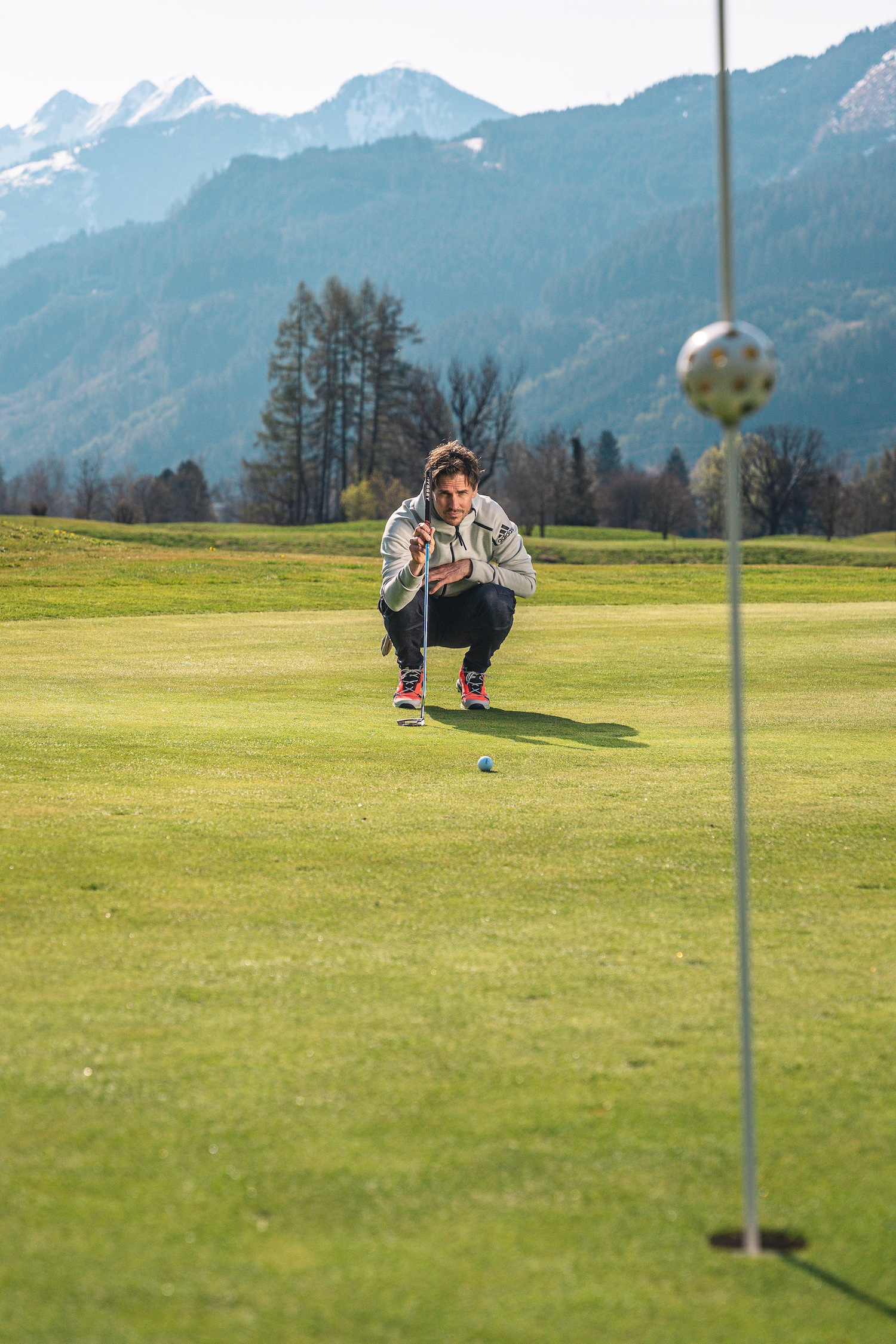 Eine Person hockt auf einem Golfplatz, um einen Putt zu spielen, mit Bergen und B&auml;umen im Hintergrund.