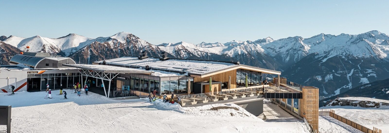 Eine moderne Skihütte mit schneebedeckten Bergen und Skifahrern im Freien unter blauem Himmel.