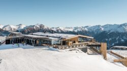 Eine moderne Skihütte mit schneebedeckten Bergen und Skifahrern im Freien unter blauem Himmel.