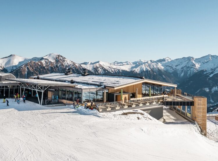 Eine moderne Skihütte mit schneebedeckten Bergen und Skifahrern im Freien unter blauem Himmel.