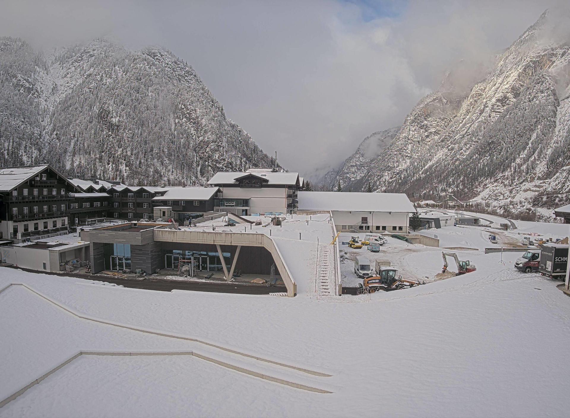 Schneebedeckte Gebäude und Fahrzeuge in einem Bergtal, umgeben von hohen, schneebedeckten Gipfeln unter einem bewölkten Himmel.