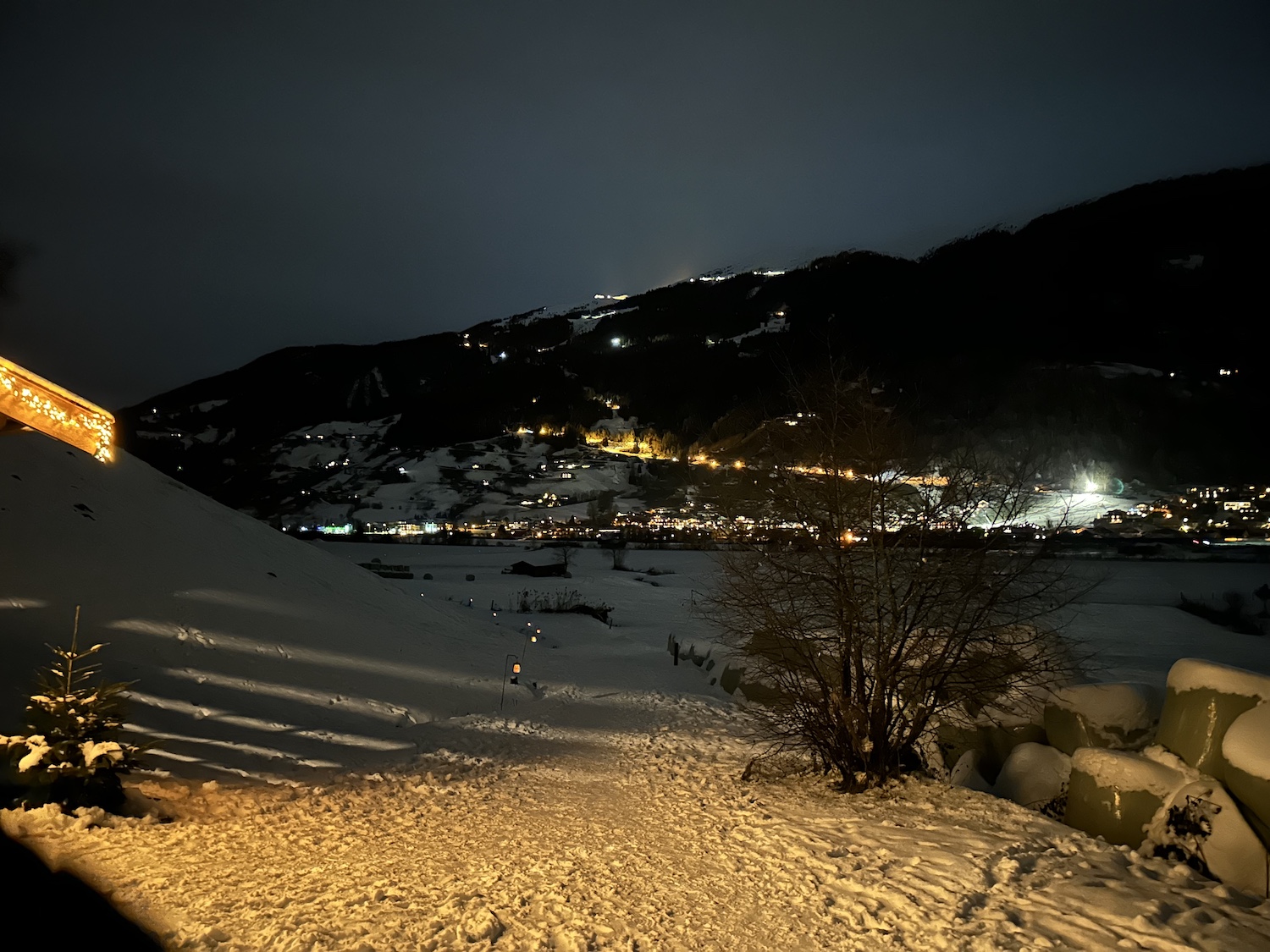 Verschneite Nachtlandschaft mit entfernten Dorflichtern am Laternenweg Bramberg, Baum und beleuchtetem Berghang.