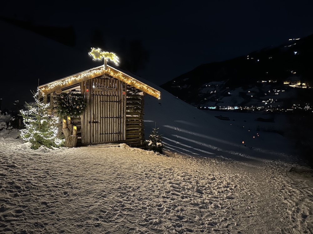 Auf dem verschneiten Laternenweg in Bramberg leuchtet nachts eine kleine Holzhütte mit Weihnachtsbeleuchtung.