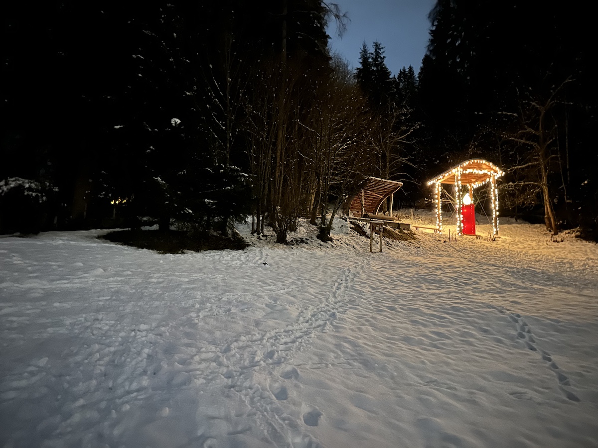 Eine verschneite Lichtung in der Abenddämmerung, ein Pavillon mit roter Tür und Lichterketten versprühen den Charme des Laternenwegs Bramberg.