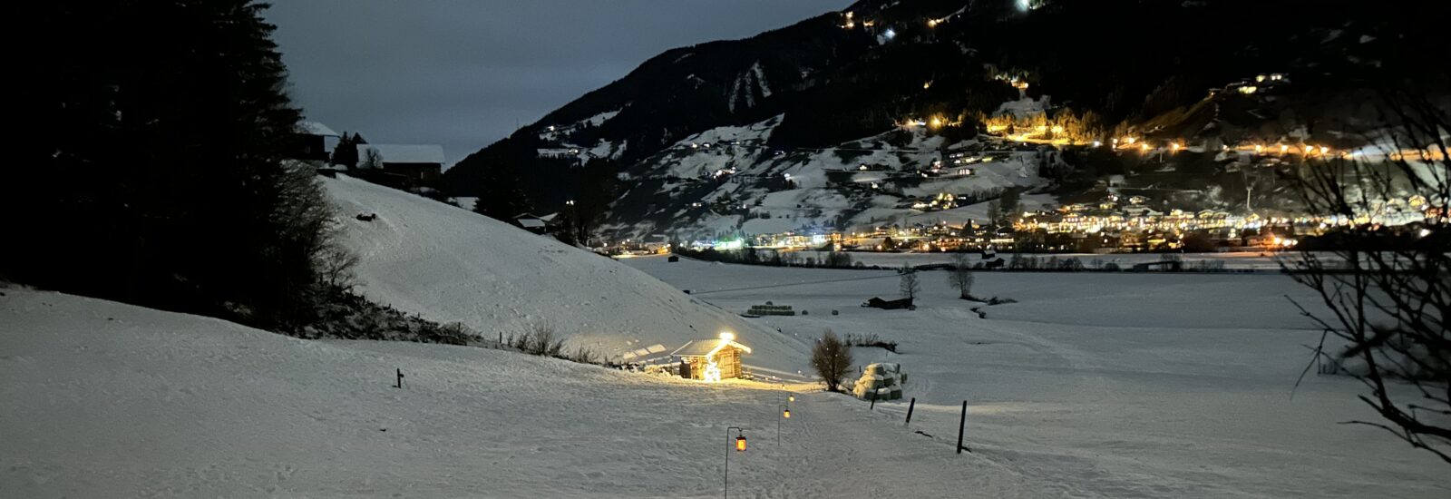 Schneebedeckte Landschaft bei Nacht mit einer beleuchteten Hütte am Laternenweg Bramberg und einer entfernten Stadt, die am Hang leuchtet.