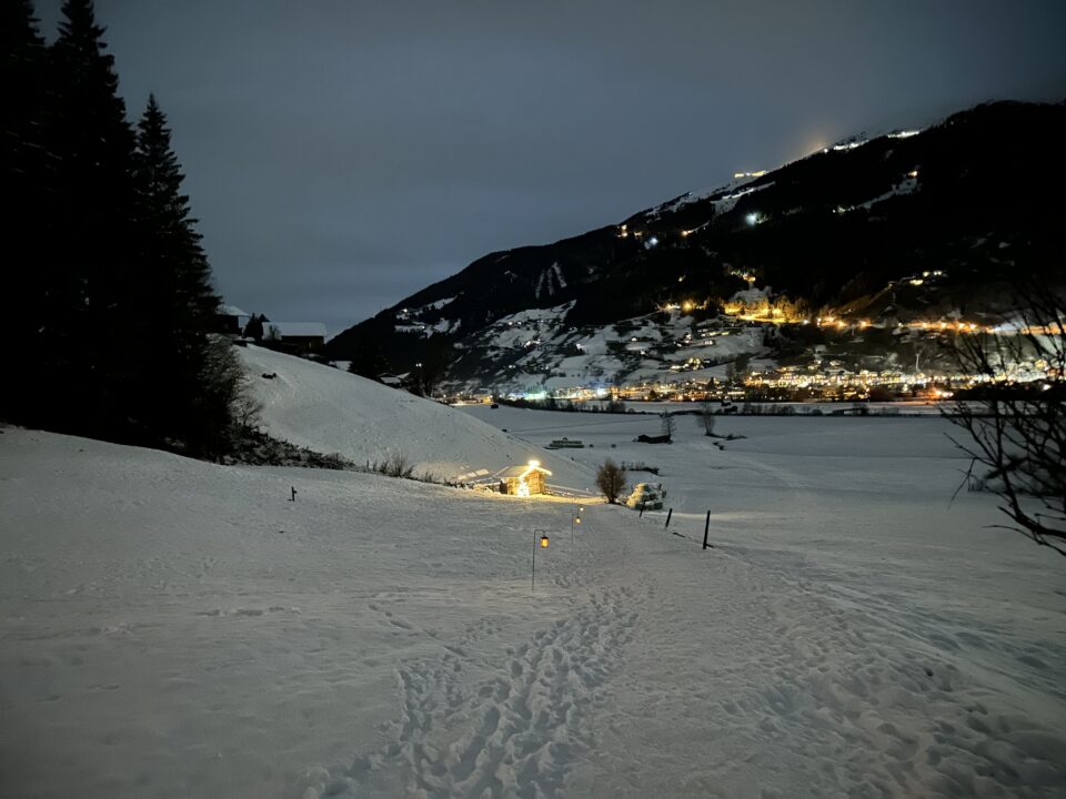Schneebedeckte Landschaft bei Nacht mit einer beleuchteten Hütte am Laternenweg Bramberg und einer entfernten Stadt, die am Hang leuchtet.