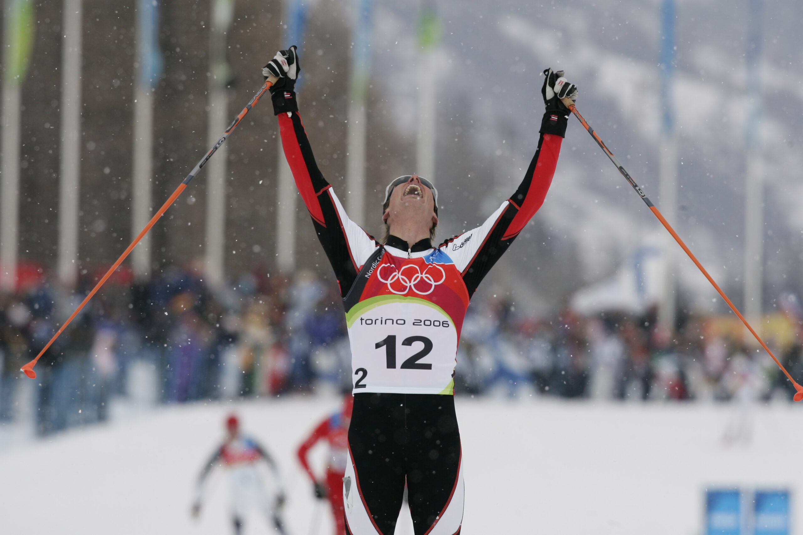 Skilangläufer in olympischer Kleidung reißt im Ziel eines verschneiten Rennens die Arme siegessicher in die Höhe, er trägt die Startnummer 12.