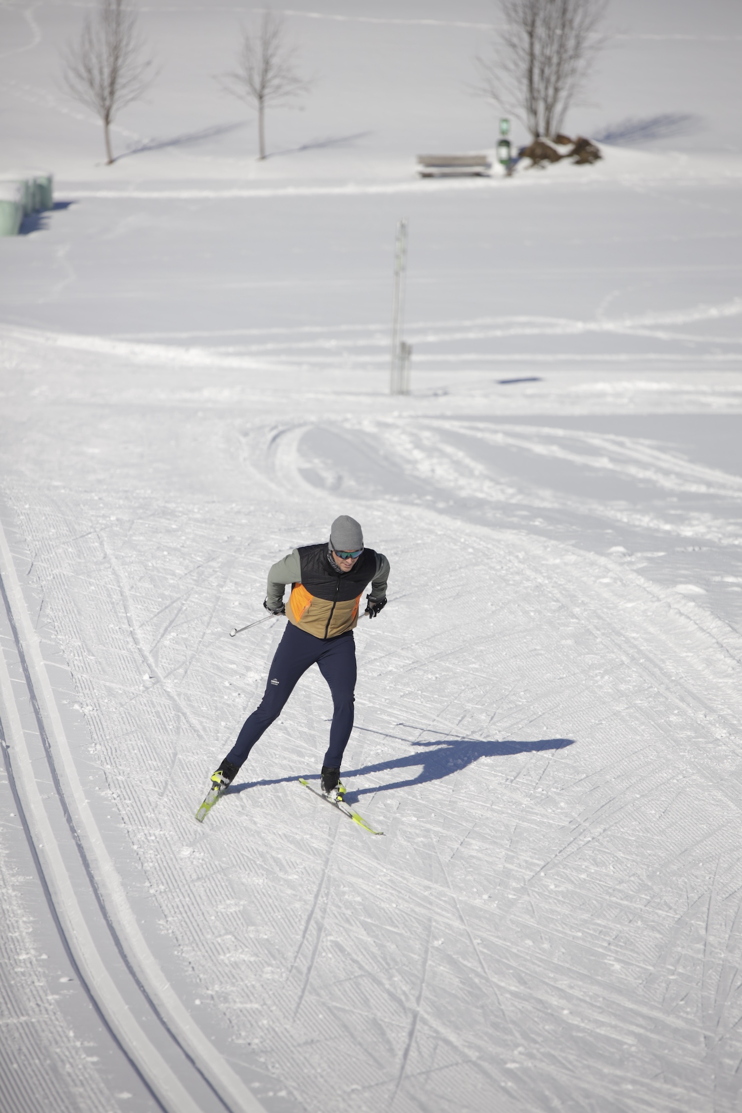 Person beim Skilanglauf auf einer verschneiten Loipe mit kahlen Bäumen im Hintergrund unter einem klaren Himmel.