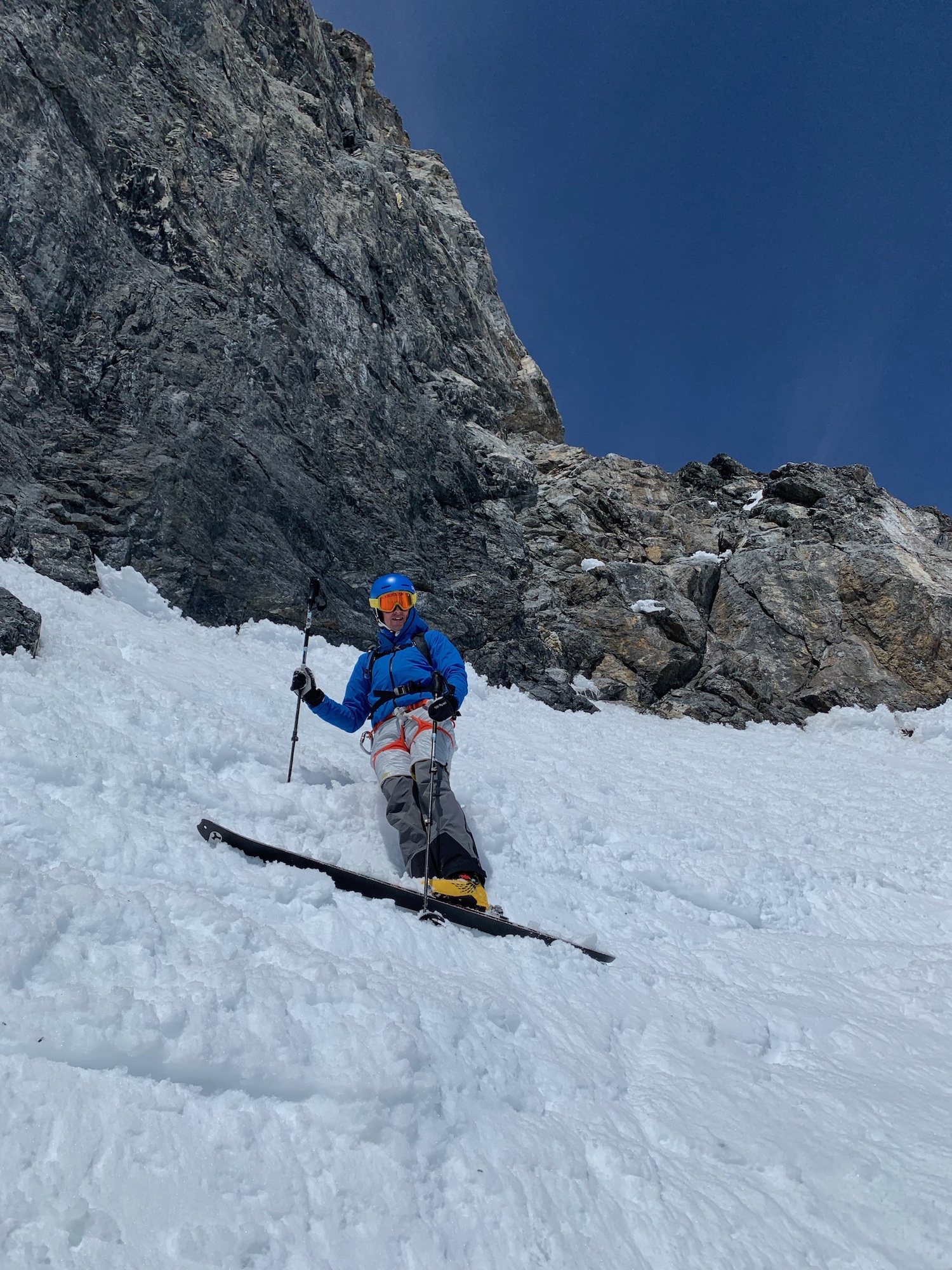 Ein Skifahrer in blauer Jacke und Helm fährt einen steilen, verschneiten Berghang in der Nähe einer felsigen Klippe unter blauem Himmel hinunter.