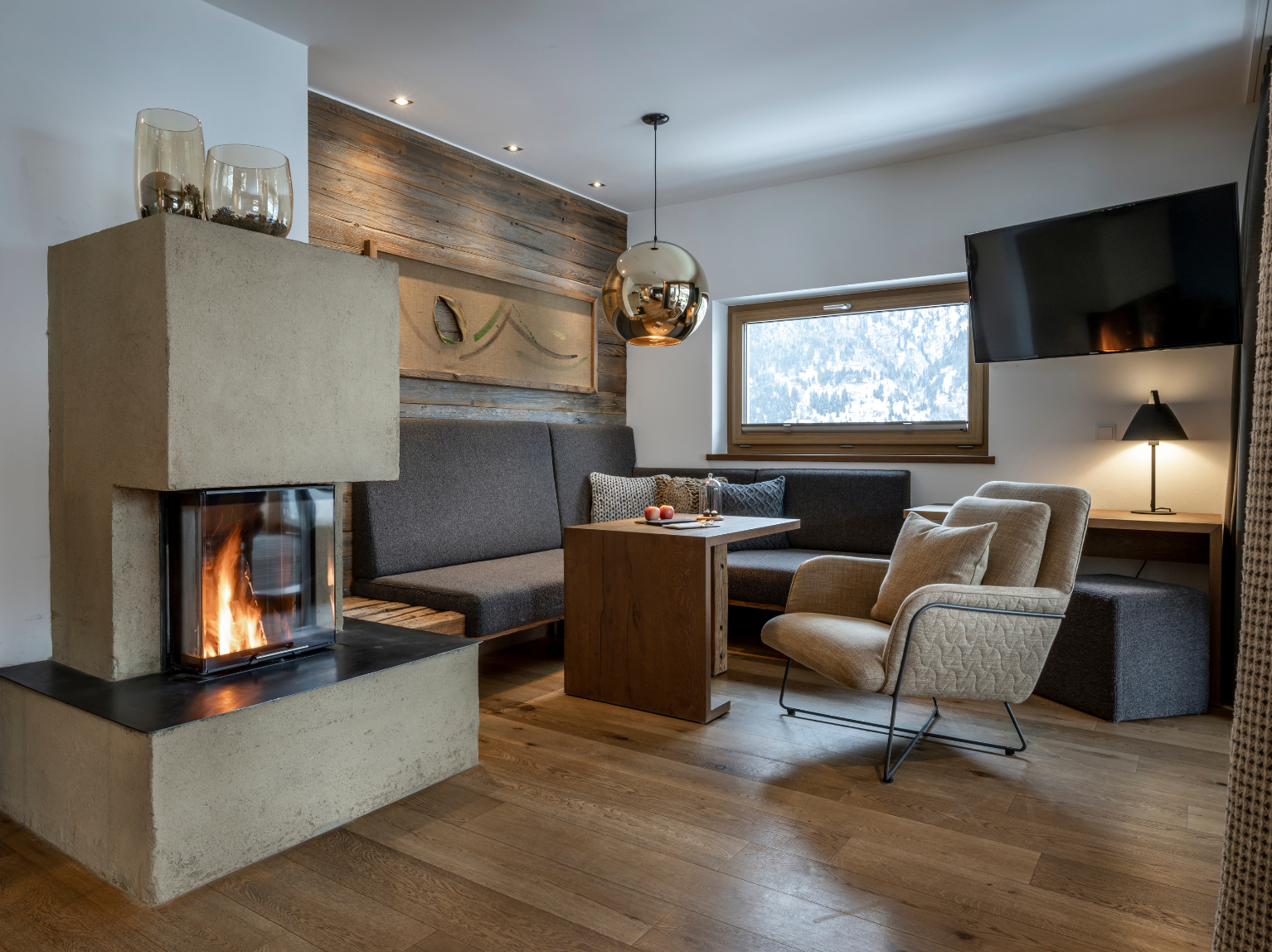 Modernes, gemütliches Wohnzimmer mit Kamin, Holzboden, Sitzecke und großem Fenster mit Blick auf die verschneiten Bäume draußen.