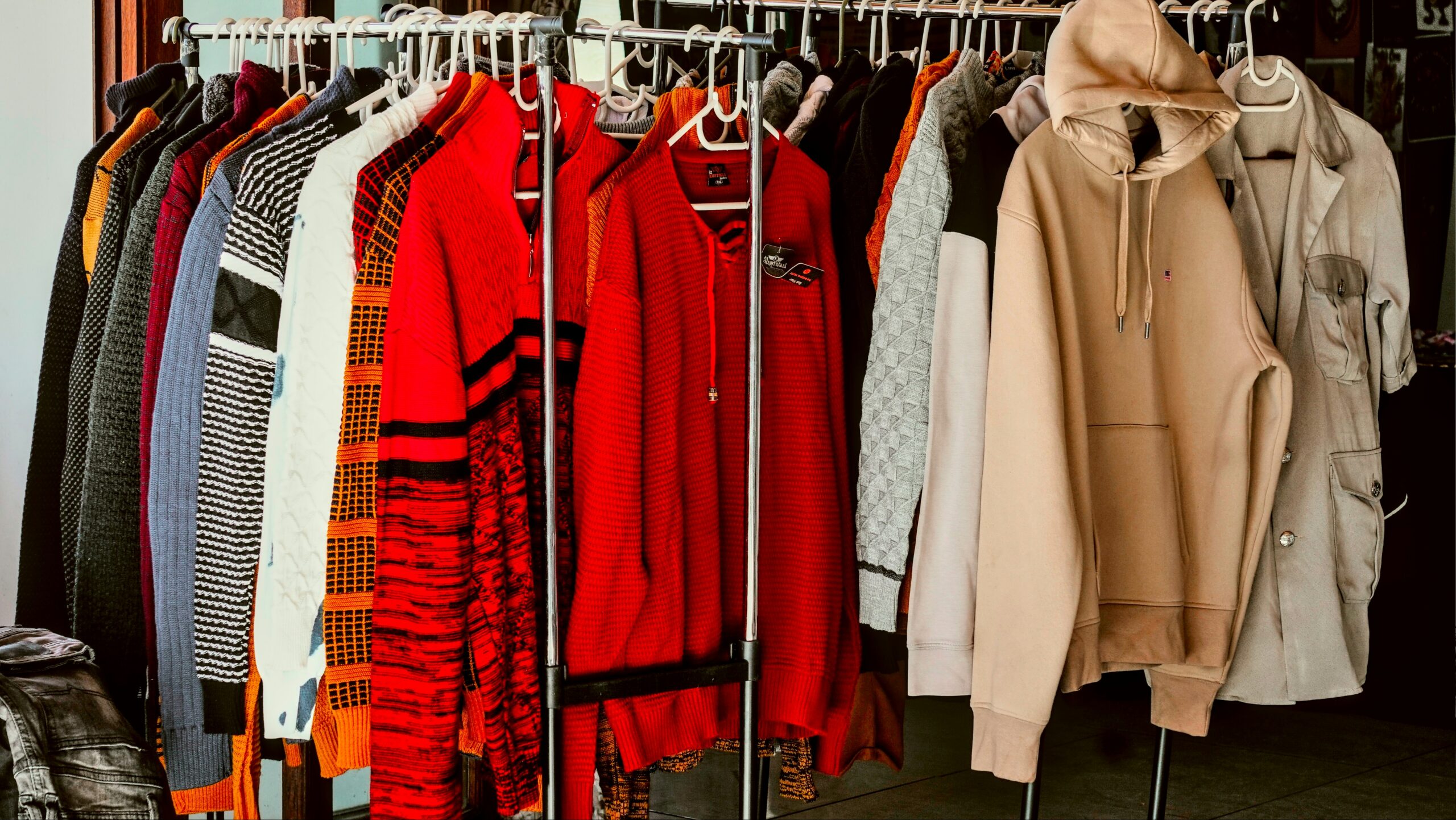 Kleiderständer mit verschiedenen bunten Pullovern, einem beigen Kapuzenpulli und einem Trenchcoat, wie man ihn bei Flohmärkten in Salzburg findet.