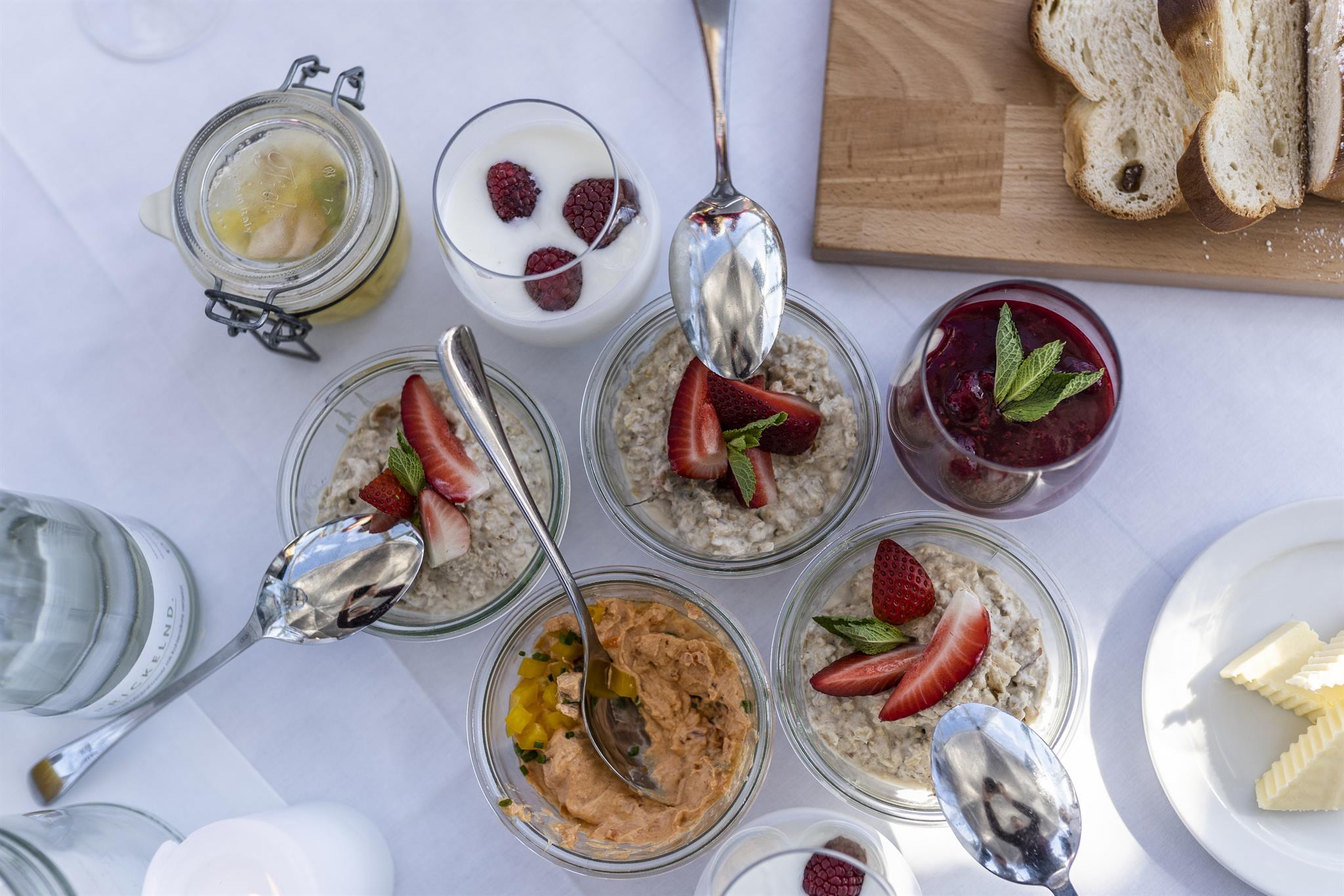 Verschiedene Joghurt- und Haferflockengl&auml;ser mit Obst, Brotscheiben und Butter auf einem wei&szlig;en Tischtuch.