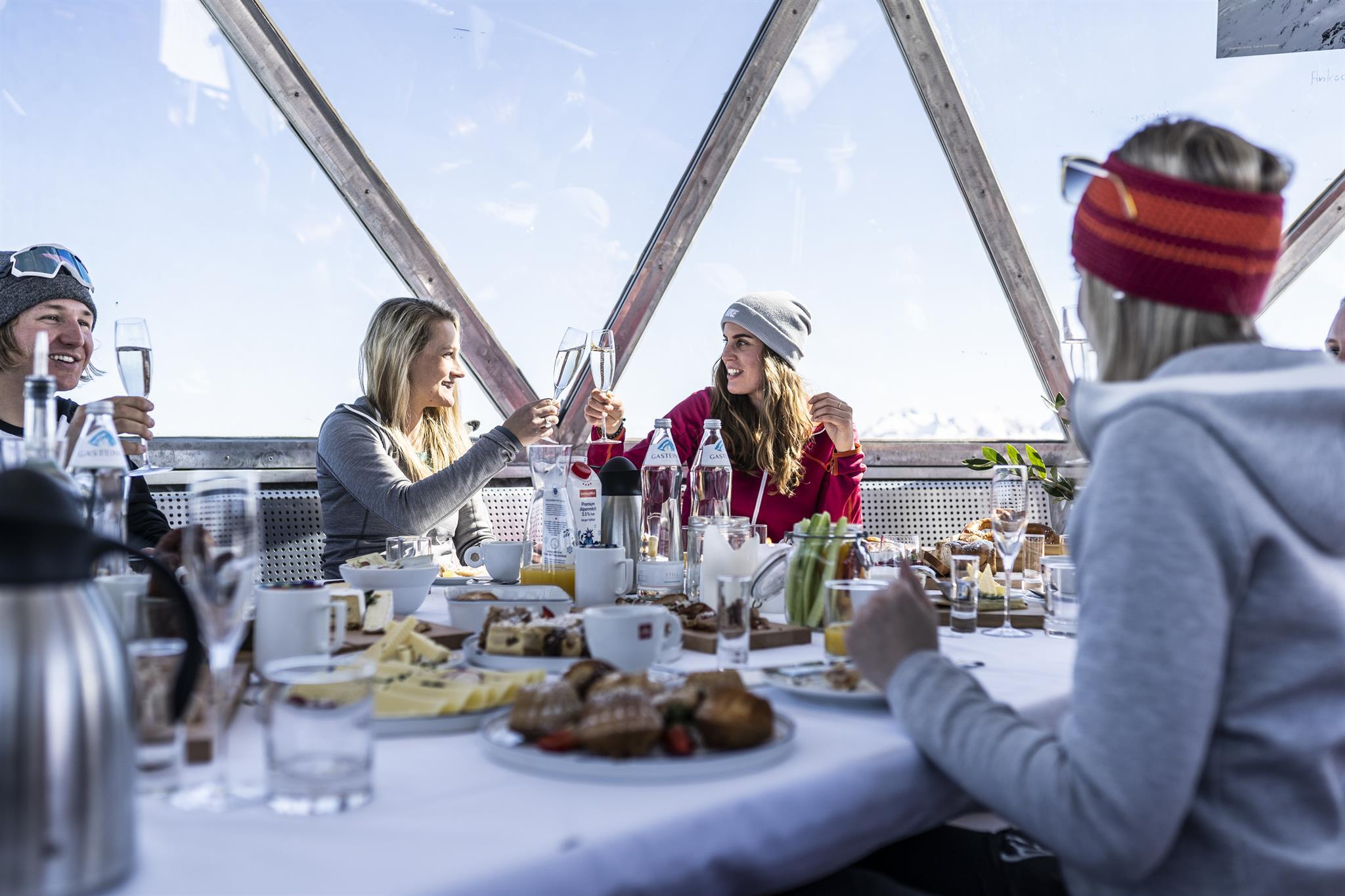 Menschen in Winterkleidung sto&szlig;en mit Getr&auml;nken um einen gedeckten Fr&uuml;hst&uuml;ckstisch in einem gl&auml;sernen Raum an.