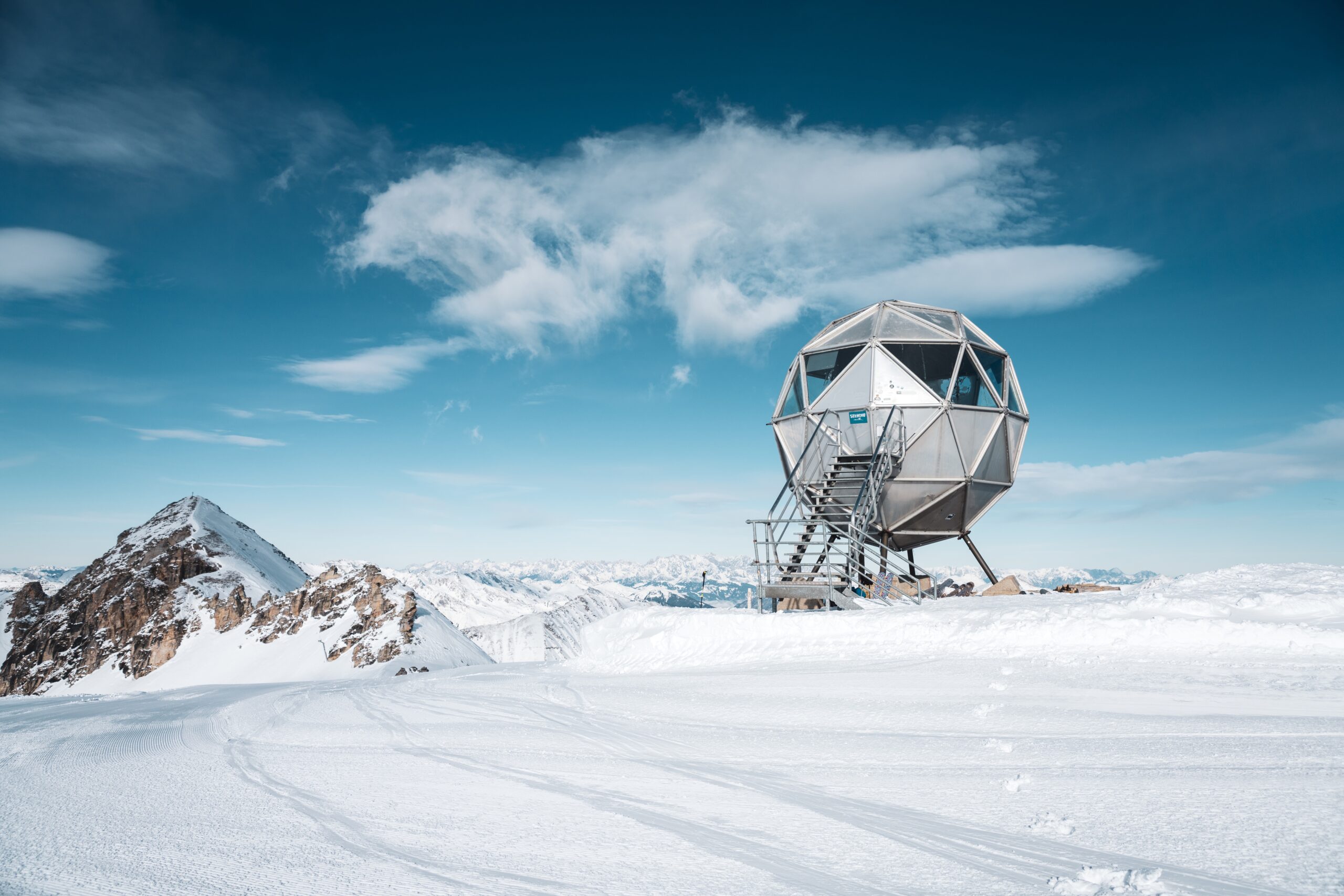 Eine geodätische Hütte auf verschneiten Bergen unter einem blauen Himmel mit vereinzelten Wolken.