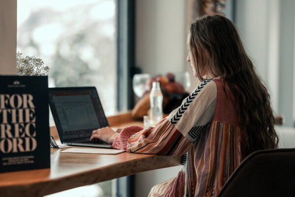 Eine Frau mit langen Haaren arbeitet an einem Laptop an einem Holztisch in der Nähe eines Fensters mit natürlichem Licht.